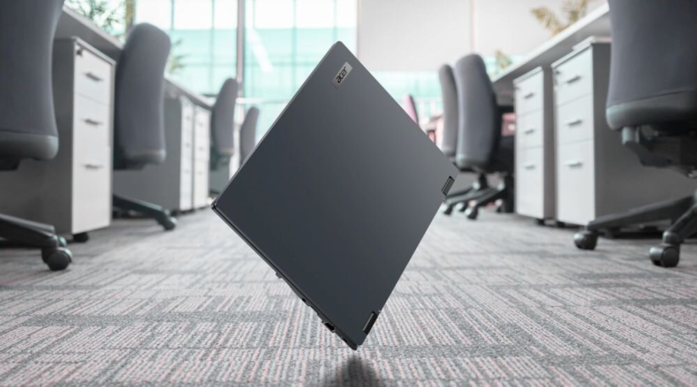 Laptop Acer TravelMate z wytrzymałą obudową unosi się nad podłogą biurową, prezentując konstrukcję do codziennej pracy biznesowej.