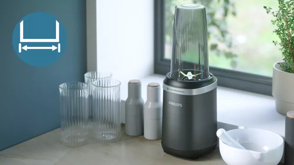 Na blacie kuchennym stoi blender kielichowy z logo PHILIPS, obok znajdują się szklanki i przyprawniki. W tle widać okno oraz roślinę doniczkową.