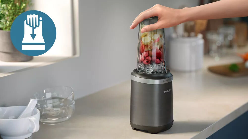 Na blacie kuchennym stoi blender kielichowy z owocami w środku, a ręka naciska na jego górną część. Obok znajduje się ikona z symbolem naciskania dłonią i strzałką w dół.