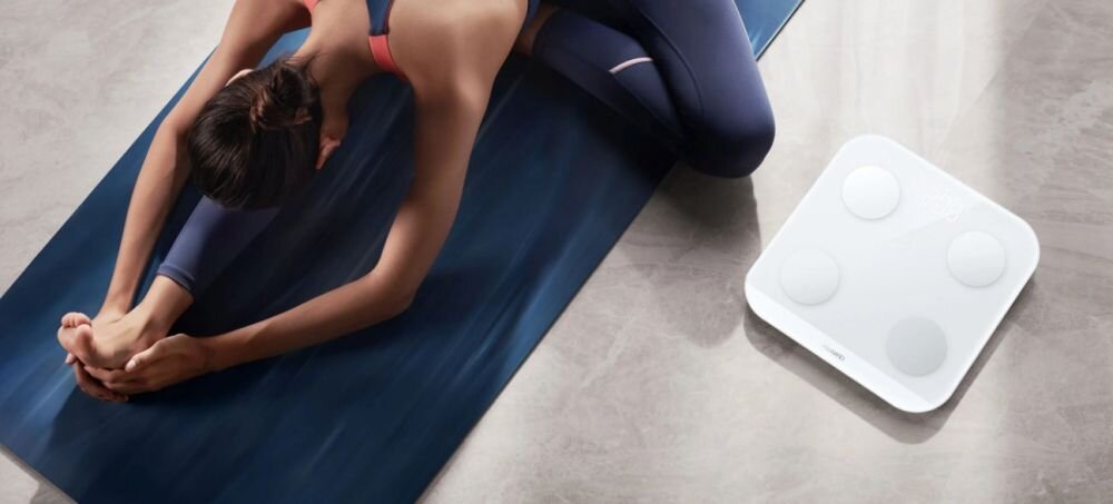 Osoba w stroju sportowym wykonuje ćwiczenia na macie. Obok znajduje się biała waga łazienkowa z czterema okrągłymi sensorami i logo HUAWEI.