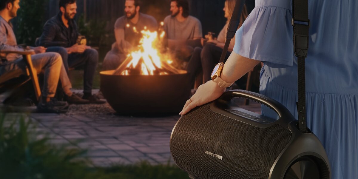 Przenośny głośnik Bluetooth Hama Ultimate Pro z paskiem na ramię, używany podczas spotkania przyjaciół przy ognisku, co podkreśla jego mobilność i zastosowanie na zewnątrz.
