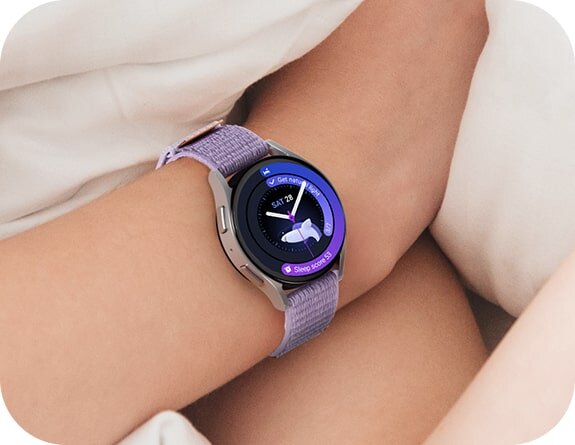 Osoba nosi smartwatch z okrągłym wyświetlaczem, na którym widoczna jest tarcza zegarka z datą \'SAT 26\' i ikoną księżyca. Pasek zegarka jest w kolorze fioletowym.