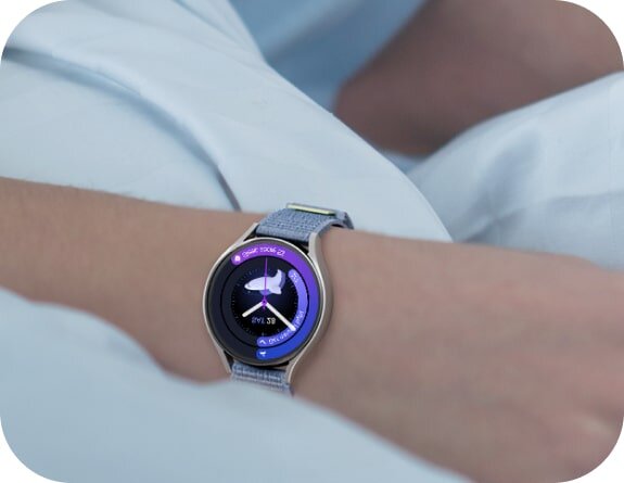 Osoba nosi srebrny smartwatch z okrągłym wyświetlaczem, na którym widoczna jest tarcza zegara z fioletowym akcentem. Pasek zegarka jest szary i teksturowany.