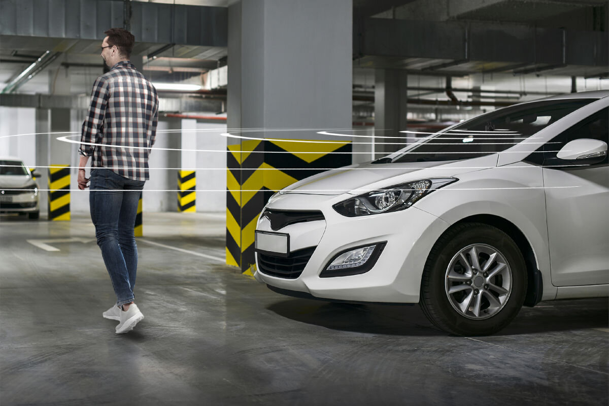 W garażu stoi biały samochód obok filaru z żółto-czarnymi pasami. Obok przechodzi osoba ubrana w koszulę w kratę i białe buty.