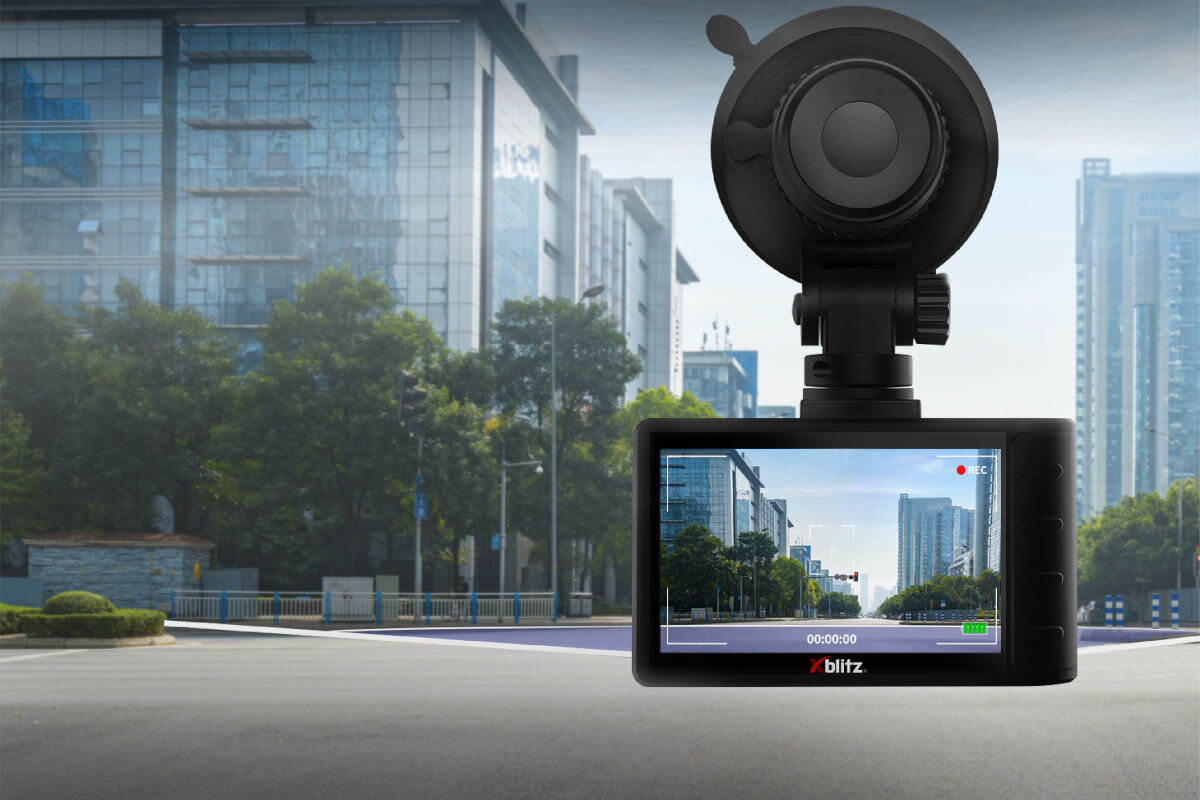 Na obrazie widoczny jest wideorejestrator z logo \'xblitz\', zamontowany na tle miejskiego krajobrazu z budynkami i drzewami. Ekran urządzenia pokazuje nagranie ulicy z podobnym widokiem.