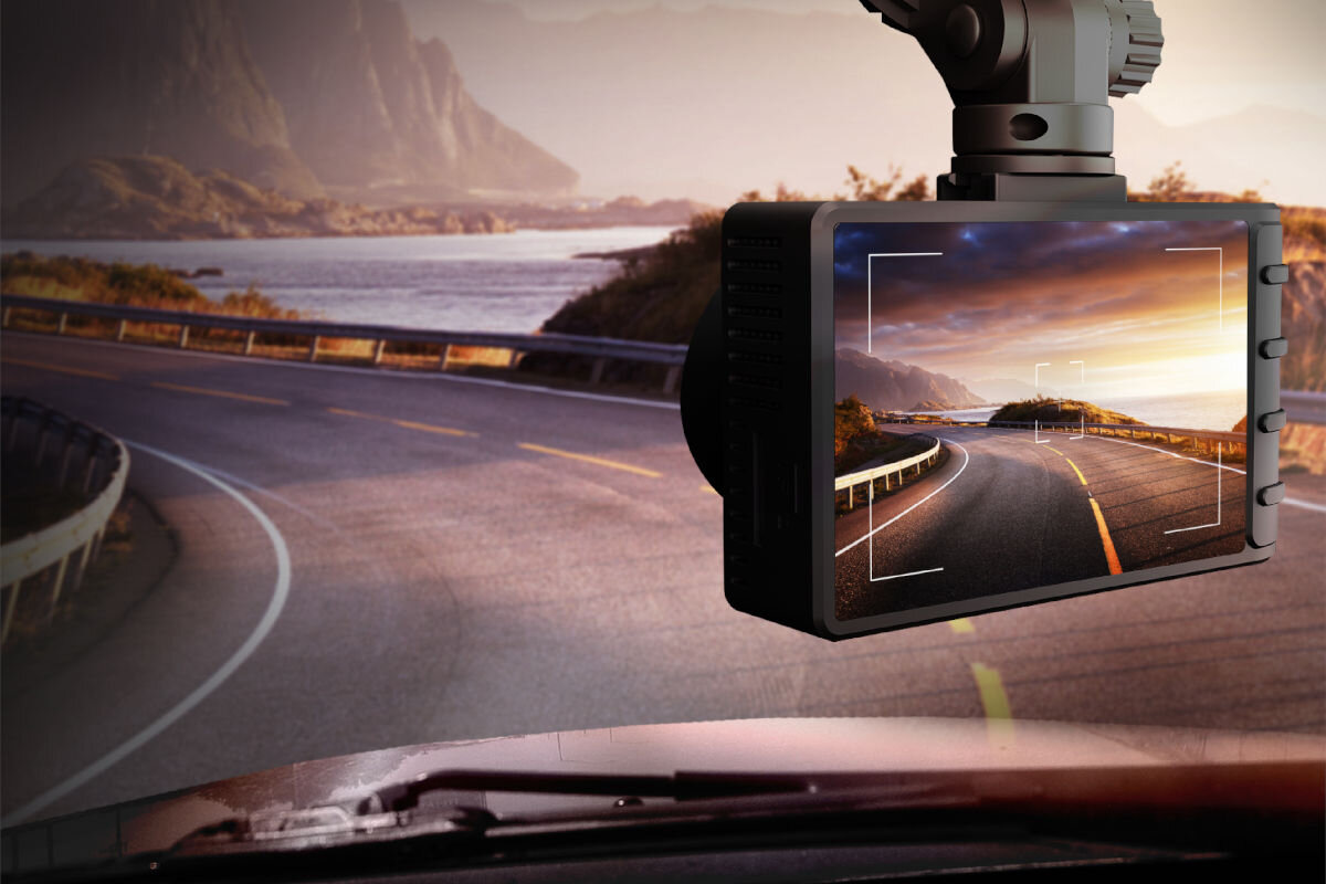 Na zdjęciu widać wideorejestrator zamontowany na przedniej szybie samochodu, który nagrywa krętą drogę z widokiem na góry i jezioro. Ekran urządzenia pokazuje tę samą scenę, co widok za szybą.