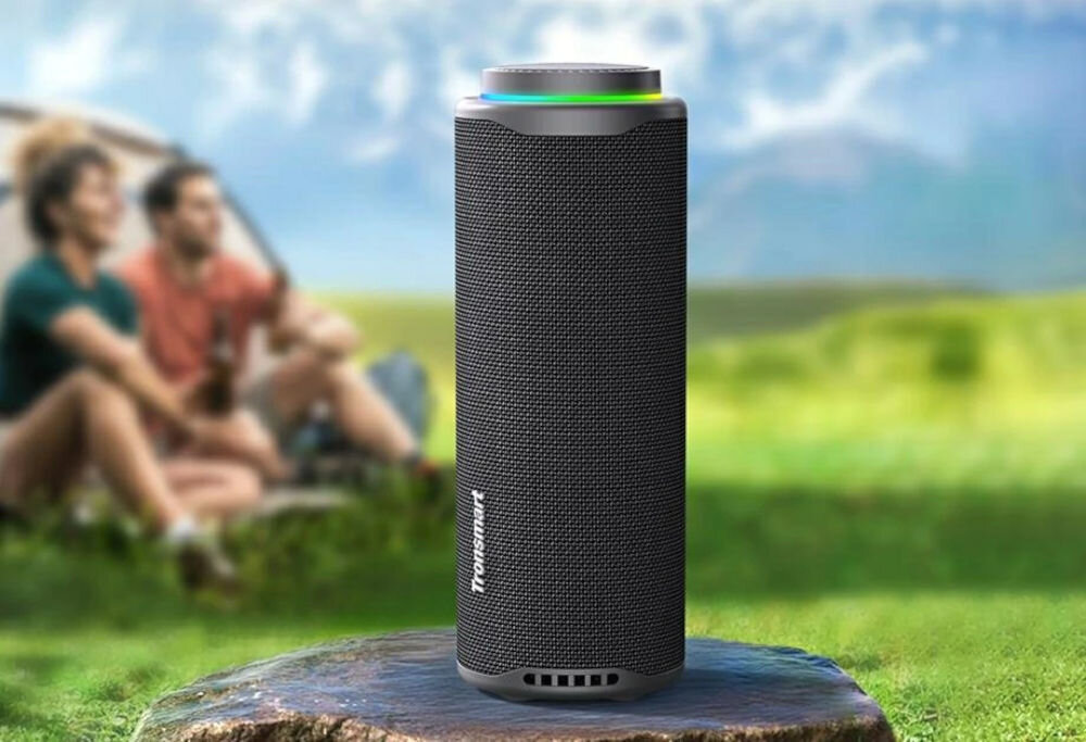 Głośnik mobilny TRONSMART T8 pełen dźwięku 360° z mocnym basem i czystymi wokalami, Głośnik T8 z logo Tronsmart ustawiony na skale na pierwszym planie, w tle dwie osoby siedzące na trawie.