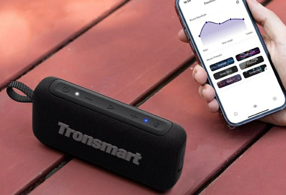 TRONSMART Trip 2 EQ aplikacja Tronsmart tryby świetlne, Głośnik Trip 2 na czerwonym drewnianym biurku, obok osoba trzymająca telefon z widocznym interfejsem aplikacji Tronsmart