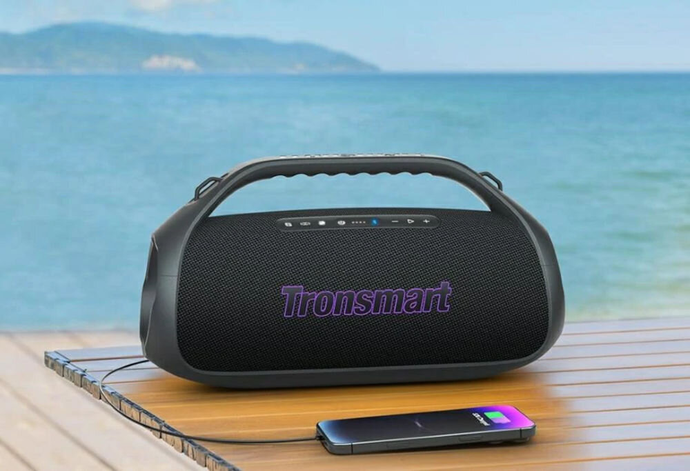 Głośnik mobilny TRONSMART Bang 2 — wbudowany powerbank, ładowanie urządzeń, mobilność, głośnik mobilny z ładującym się smartfonem na drewnianym stole na tle oceanu