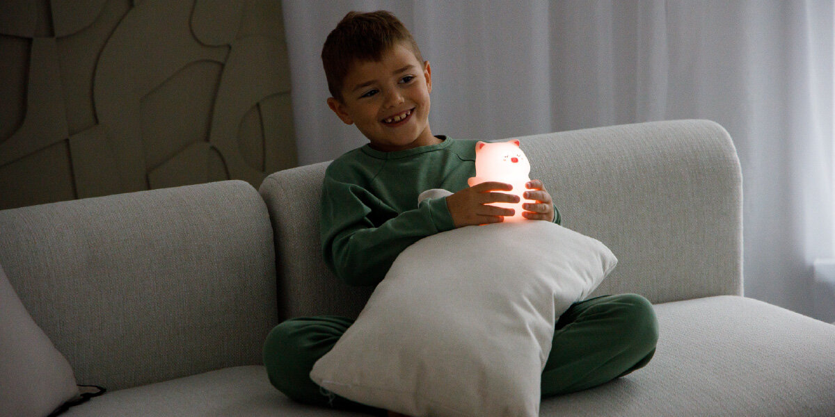 Lampka nocna INNOGIO GIOsleepy Świnka GIO-144, bezpieczna, dotyk, przytulanie, BPA, dziecko siedz na sofie trzymając lampkę nocną przy poduszcze