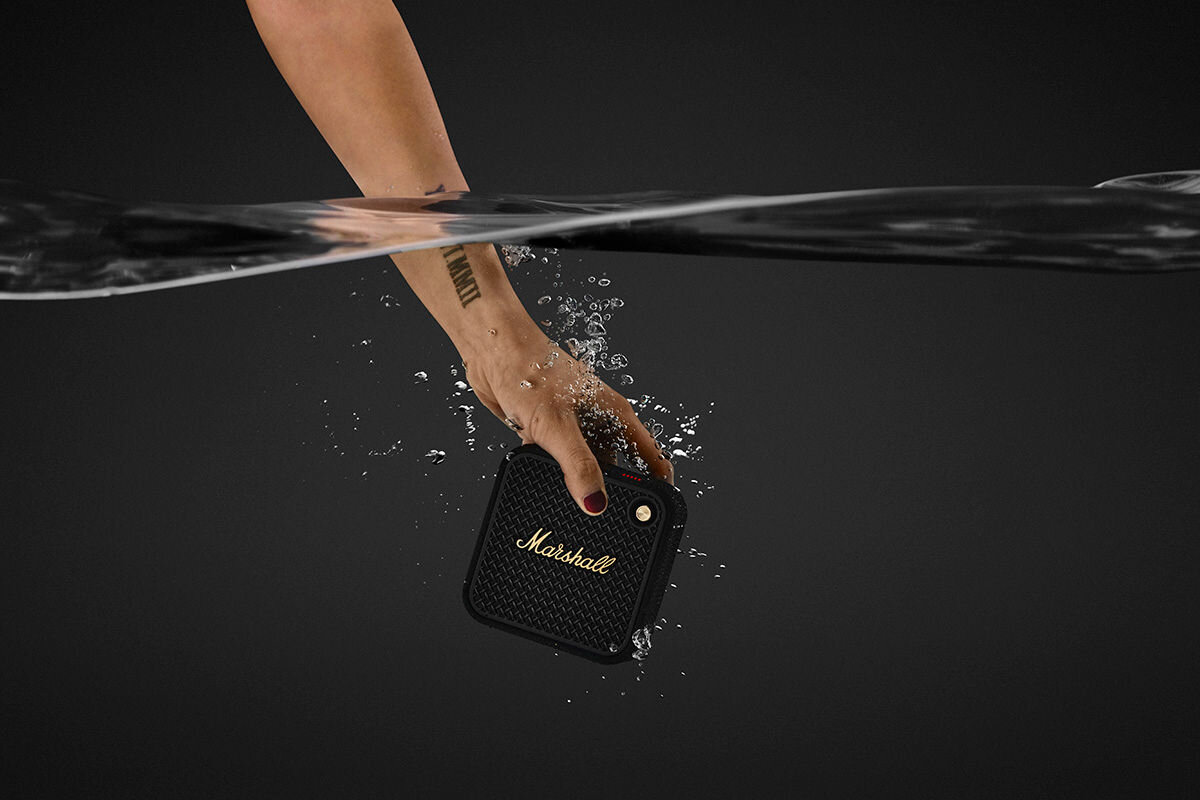 Ręka zanurza czarno-miedziany głośnik mobilny z logo \'Marshall\' w wodzie. Widać rozpryski wody wokół głośnika.