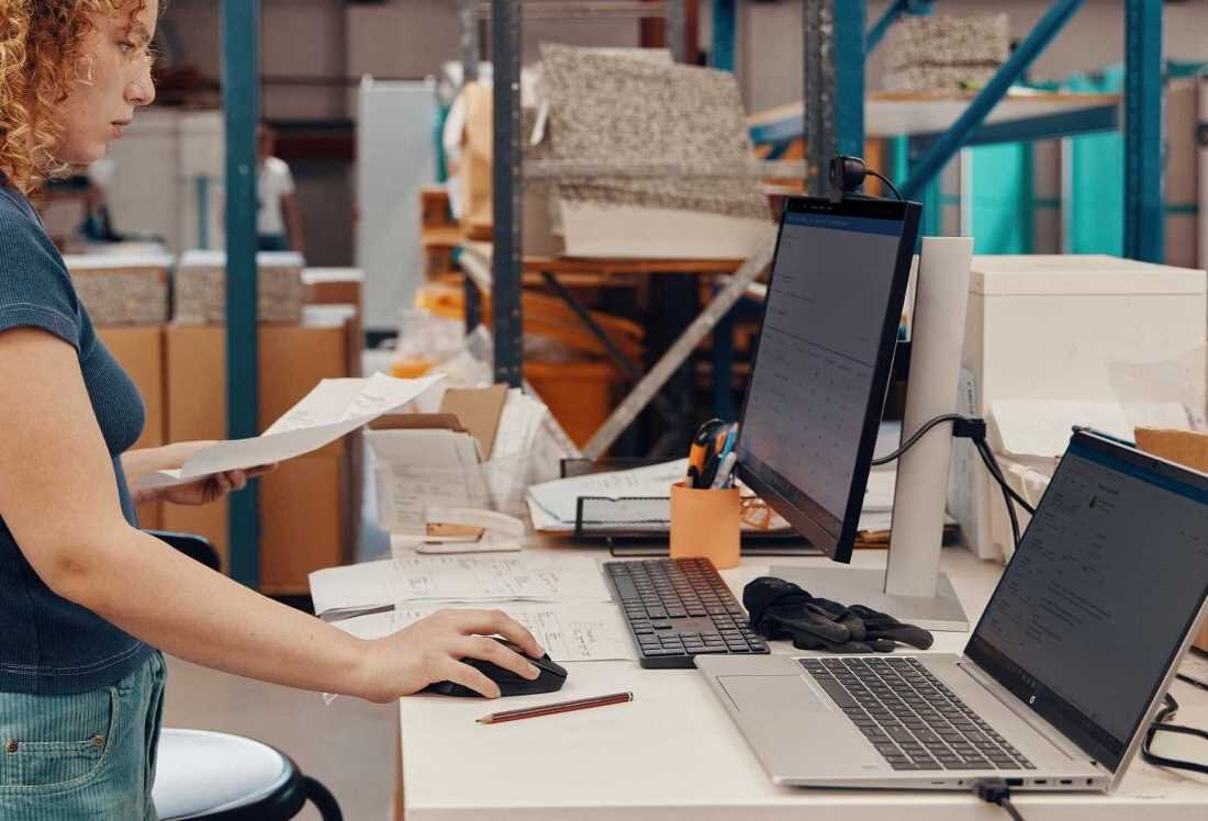 Kobieta w niebieskiej koszulce z kręconymi włosami pracuje przy biurku w magazynie, używając laptopa HP ProBook 450 G10, zewnętrznego monitora i myszki, przeglądając dokumenty.   