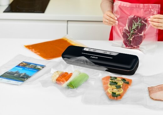 Zgrzewarka  MPZ-02na kuchennym blacie. Dookoła woreczki próżniowe z żywnością. W tle kobieta trzyma woreczek z mięsem.