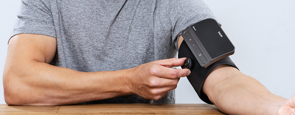 Osoba siedzi przy stole, używając ciśnieniomierza na ramieniu. Na urządzeniu widoczne jest logo \'Beurer\'.