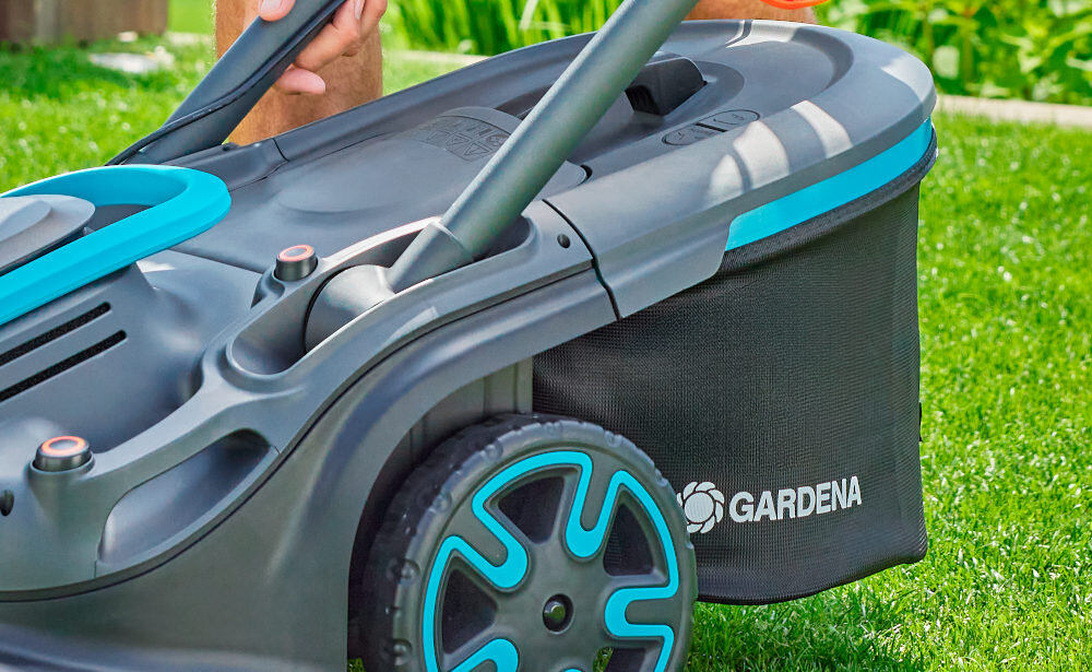 Na obrazie widoczna jest kosiarka akumulatorowa z logo \'GARDENA\' na czarnym koszu. Kosiarka ma szare i niebieskie elementy oraz duże koła z niebieskimi akcentami.