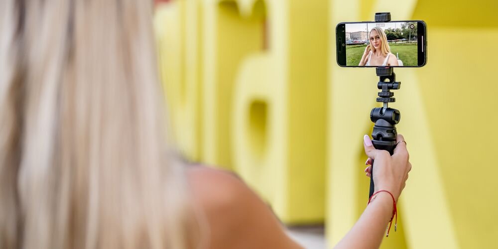 Osoba trzyma smartfon zamocowany na uchwycie, na ekranie widoczna jest jej twarz. W tle znajdują się duże, żółte litery.