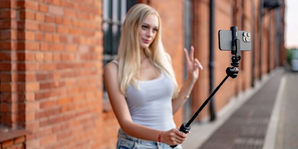 Osoba trzymająca kijek do selfie z zamocowanym smartfonem na tle ceglanej ściany. Osoba wykonuje gest ręką, a w tle widać chodnik i budynek.
