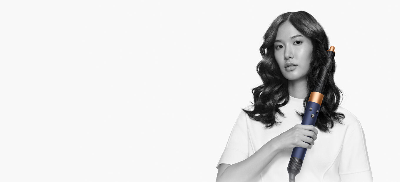 Osoba trzyma suszarko-lokówkę o nowoczesnym designie z ciemnym uchwytem i jasną końcówką. Tło jest jednolite i jasne.