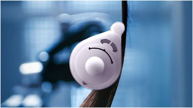 Na obrazie widoczna jest suszarko-lokówka z białą końcówką, która stylizuje włosy. W tle znajduje się rozmyte, niebieskawe tło.