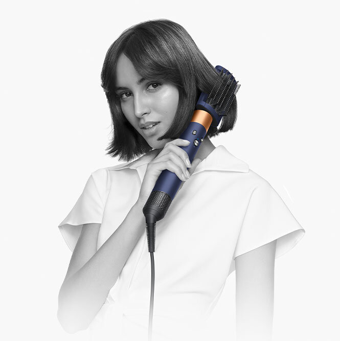 Osoba trzyma suszarko-lokówkę o ciemnym kolorze z miedzianymi elementami, przyłożoną do włosów. Ubrana jest w jasną bluzkę z kołnierzykiem.