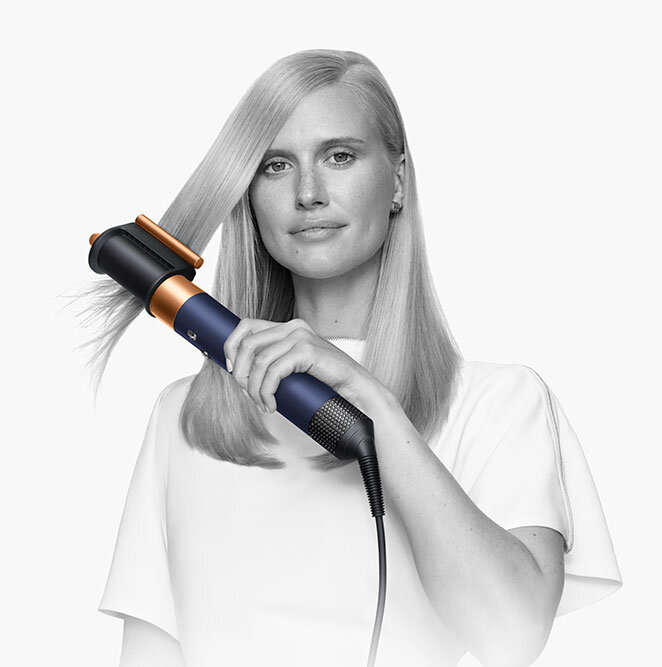 Osoba używa suszarko-lokówki o nowoczesnym designie, z elementami w kolorze niebieskim i pomarańczowym. W tle jednolite, jasne tło.