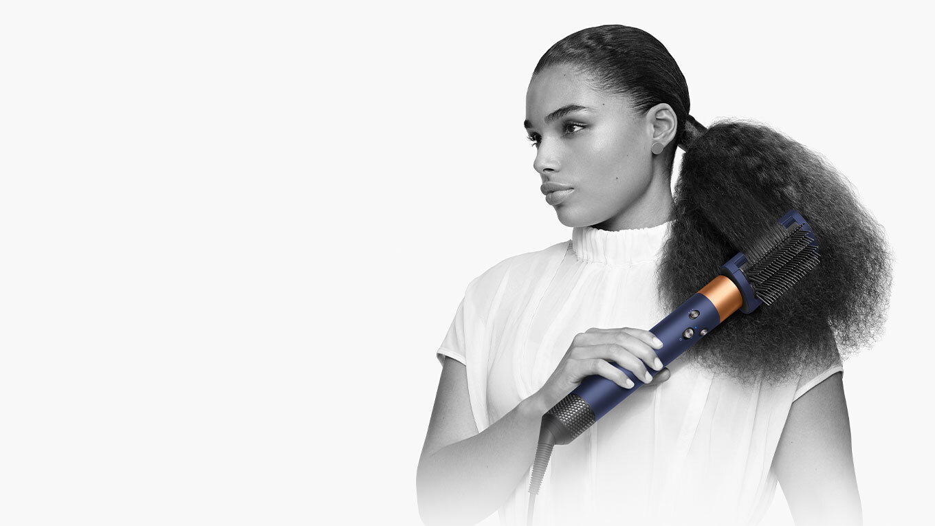 Osoba używa suszarko-lokówki o ciemnym kolorze z widocznymi elementami w kolorze miedzianym. W tle jednolite, jasne tło.