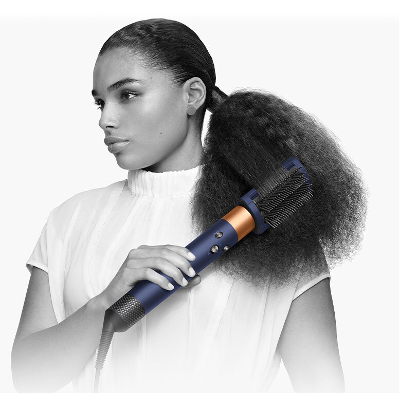 Osoba używa suszarko-lokówki o ciemnym kolorze z metalicznymi elementami do stylizacji włosów. W tle jednolite, jasne tło.