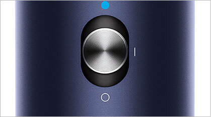 Zbliżenie na okrągły, metaliczny przycisk z teksturą, otoczony czarnym obramowaniem na ciemnoniebieskim tle. Nad przyciskiem znajduje się mała, niebieska kropka, a poniżej biały okrąg.