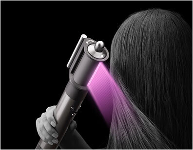 Osoba używa urządzenia do stylizacji włosów, które emituje różowe światło na włosy. Tło jest czarne, a urządzenie ma metaliczny wygląd.