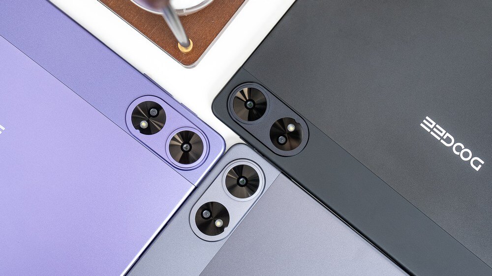 Na obrazie widoczne są trzy tablety w różnych kolorach: fioletowym, szarym i czarnym, z widocznymi aparatami fotograficznymi. Na czarnym tablecie znajduje się logo \'DOOGEE\'.