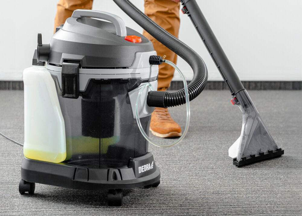 Odkurzacz piorący DEDRA DED6608 pomieszczenie osoba odkurza dywan w tle biała ściana funkcja wydmuchu powietrz skuteczne czyszczenie trudno dostępnych miejsc usuwanie nagromadzonego kurzu i pyłu