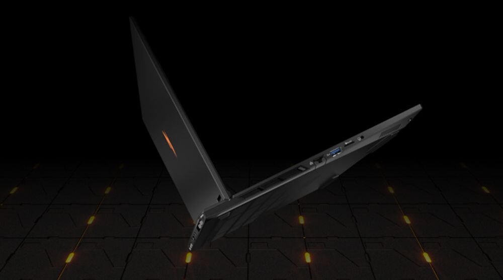 Laptop w kolorze czarnym, częściowo otwarty, widoczny z boku na tle ciemnej powierzchni z pomarańczowymi akcentami. Na obudowie znajduje się pomarańczowe logo.