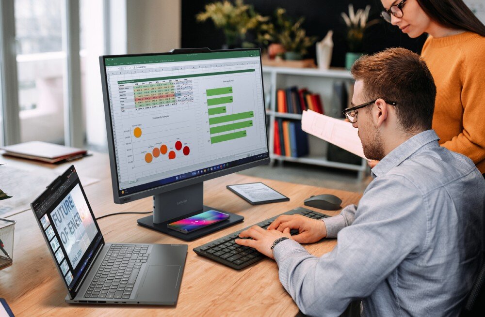 Komputer All in One LENOVO ThinkCentre neo 50a 27 Gen 5 Mężczyzna w okularach pracuje przy drewnianym biurku, pisząc na czarnej klawiaturze, przed nim stoi duży komputer z arkuszem kalkulacyjnym na ekranie, obok leży smartfon na podstawce, otwarty laptop oraz mały tablet, opis