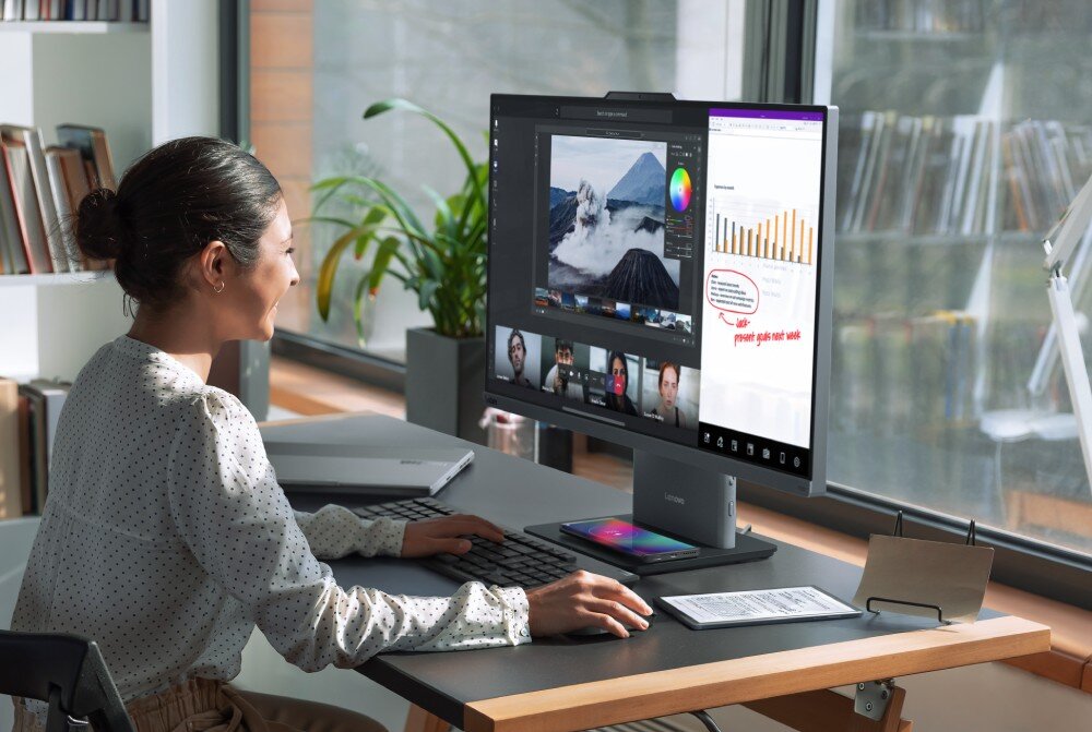 Komputer All in One LENOVO ThinkCentre neo 50a 27 Gen 5 Kobieta z upiętymi włosami siedzi bokiem przy szarym biurku, obsługując myszką i klawiaturą komputer, na którego dużym ekranie wyświetla się program do edycji zdjęć ze spotkaniem online oraz arkusz z notatkami, dysk, pojemność, Copilot