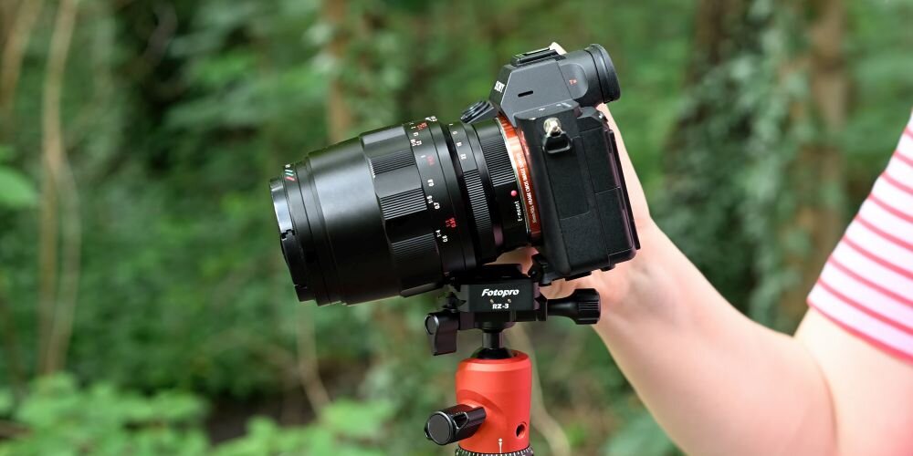 Osoba trzyma aparat fotograficzny zamontowany na statywie z adapterem Fotopro RZ-3. W tle widoczna jest zielona roślinność.