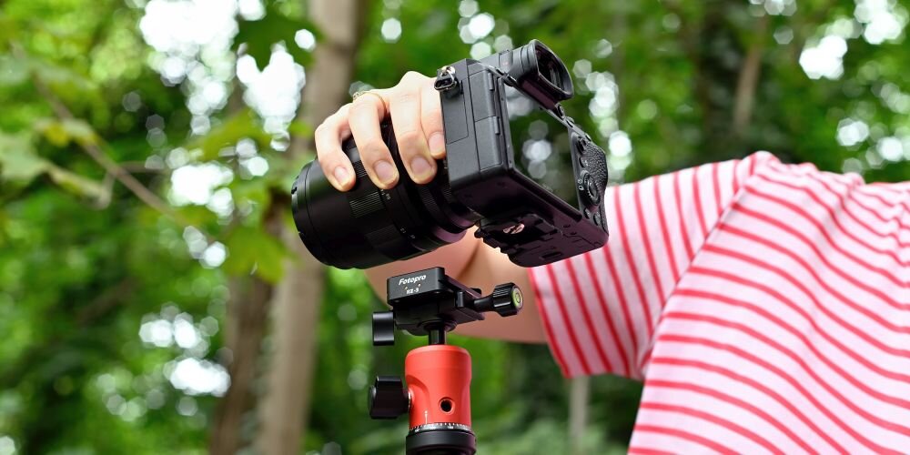 Osoba w czerwono-białej koszulce trzyma aparat zamontowany na statywie z adapterem Fotopro RZ-3. W tle widoczne są zielone drzewa.