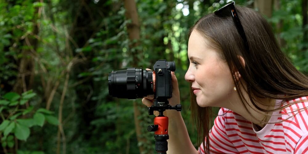 Osoba trzyma aparat fotograficzny zamontowany na statywie w otoczeniu zieleni. Statyw posiada czarny adapter z widocznym napisem \'Fotopro\'.