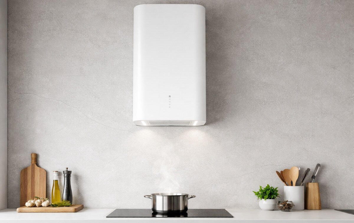 Na obrazie widoczny jest biały okap kuchenny zamontowany nad płytą grzewczą, na której stoi garnek z parą. W tle znajduje się szara ściana oraz kilka akcesoriów kuchennych, takich jak deska do krojenia i butelki z oliwą.