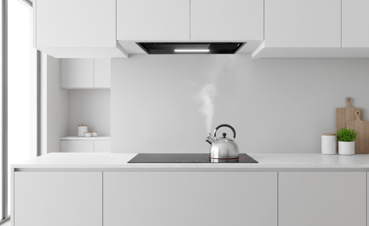 OKAP AKPO WK-9 CLARA CZARNY T500 Zdjęcie przedstawia minimalistyczną kuchnię w jasnych barwach, z płytą grzewczą umieszczoną na wyspie i czajnikiem, z którego unosi się para. Nad płytą wbudowany jest czarny okap sufitowy z oświetleniem LED, podkreślający czystą i prostą formę wnętrza