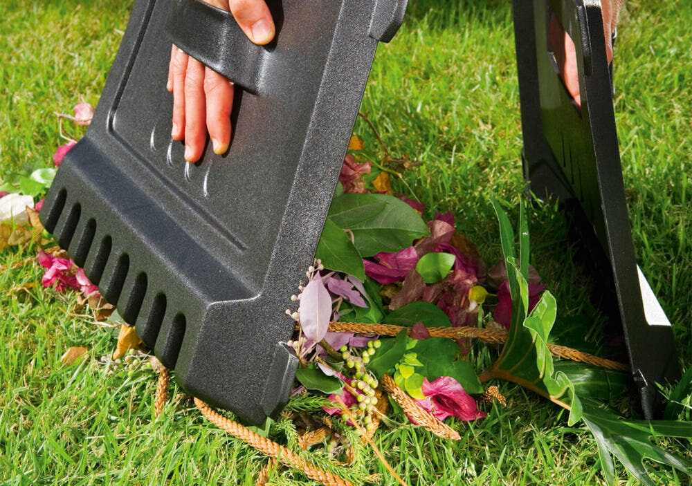 Dwie ręce trzymają czarne plastikowe elementy nad stosikiem liści i kwiatów na trawie. Elementy mają ząbkowane krawędzie i uchwyty na dłonie.