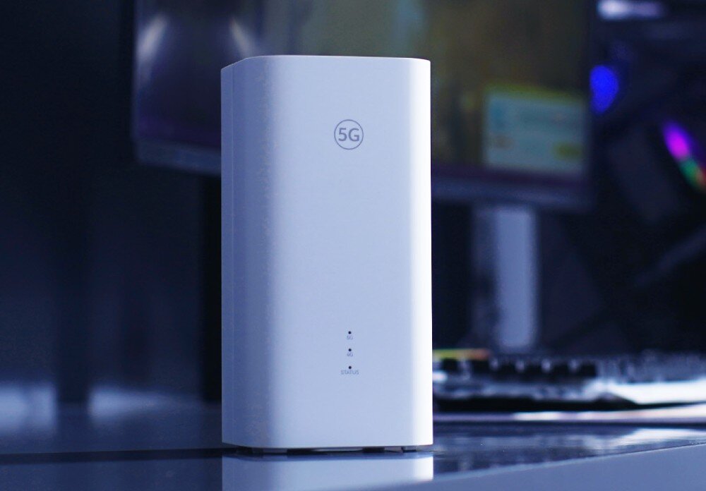 Router HUAWEI Zowee 5G router stojący na ciemnym blacie w pomieszczeniu o chłodnym oświetleniu, w tle widoczny jest monitor komputerowy