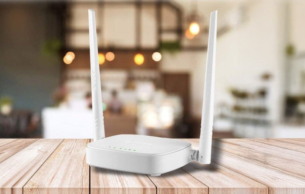 Router TENDA N301 2.4 GHz Mały biały router z dwiema antenami ustawiony na drewnianym stole. W tle rozmyte wnętrze kawiarni lub restauracji z ciepłym oświetleniem, opis