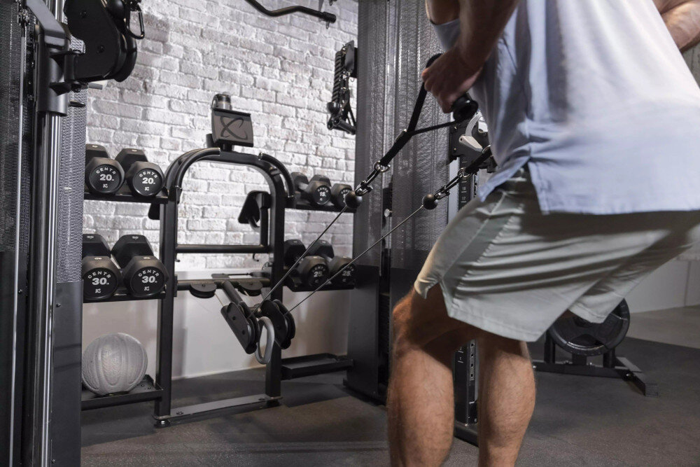 Brama do ćwiczeń INSPIRE FITNESS FT2 PRO + Ławka FID7.1 pomieszczenie siłownia brama na tle ceglanej ściany osoba ćwiczy ramiona system sześciu bloczków regulowany bloczek środkowy szeroki zakres ruchów wykonywanie różnych ćwiczeń użytkownik może stopniowo rozwijać mobilność i siłę płynny przesuw linek