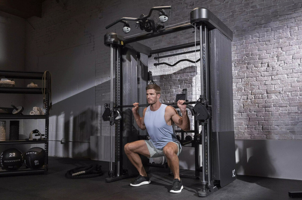 Brama do ćwiczeń INSPIRE FITNESS FT2 PRO + Ławka FID7.1 pomieszczenie siłownia brama na tle ceglanej ściany osoba ćwiczy przysiady suwnica Smitha przysiady wyciskania i inne ćwiczenia z prowadzeniem stabilność ruchu redukuje ryzyko urazu maksymalne obciążenie do 208,7 kg stałe prowadzenie gryfu poprawna technika