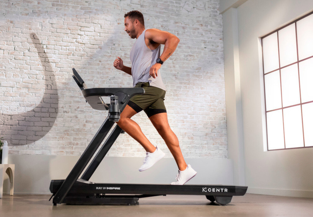 Bieżnia elektryczna CENTR RunR-S 2238W 16km-h pomieszczenie bieżnia na środku pomieszczenia osoba biegnie na bieżni w tle okno ściana z cegły poziom hałasu 56 dB przy 10 km/h komfortowy trening bez zakłócania spokoju domowników stabilna konstrukcja szeroki pas amortyzacja komfortowa w codziennym użytkowaniu