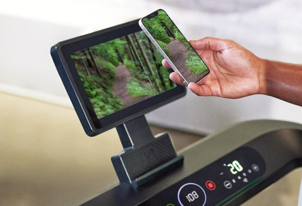 Bieżnia elektryczna CENTR RunR-S 2238W 16km-h zbliżenie na ekran osoba trzyma telefon w dłoni ze sklonowanym ekranem 10-calowy ekran dotykowy 25 programów treningowych programy HRC interwałowe kontrola tętna i intensywności wysiłku funkcja mirroringu klonowanie ekranu smartfona lub tabletu na ekranie bieżni bezpośrednio na ekranie bieżni komfort i motywacja