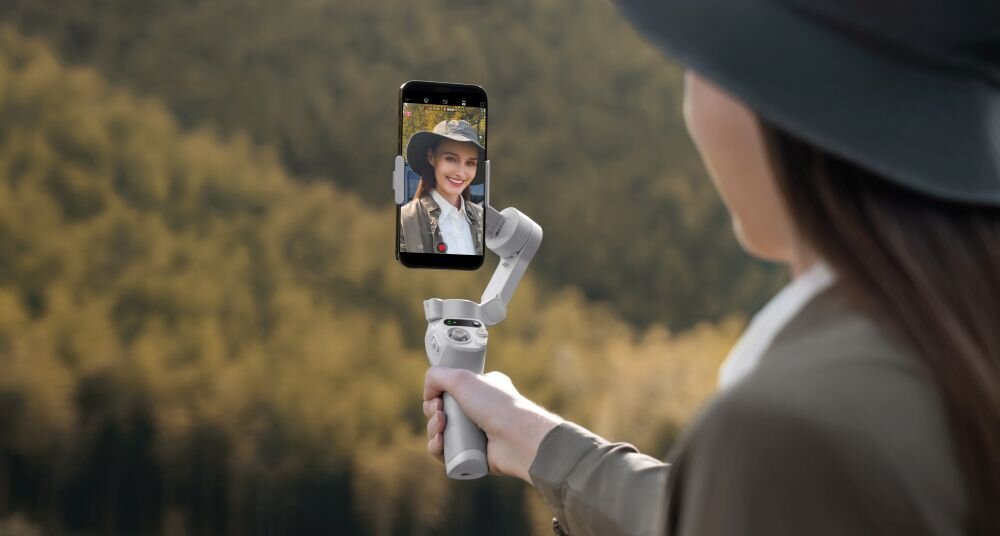 Osoba trzyma gimbal z zamocowanym smartfonem, na którym widać uśmiechniętą kobietę. W tle znajduje się rozmyty krajobraz z drzewami.