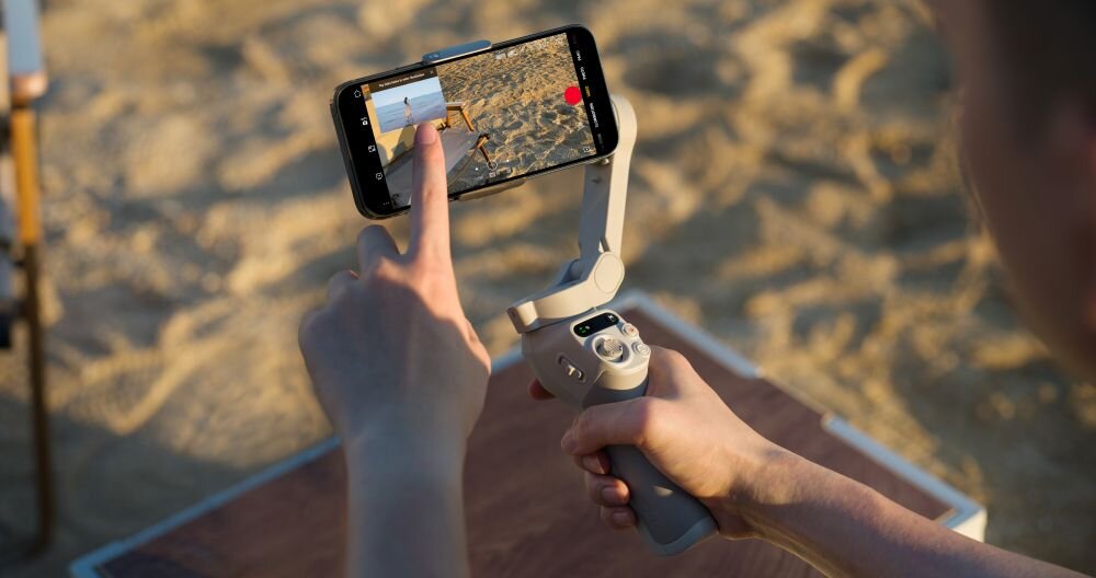 Osoba trzyma gimbal z zamocowanym smartfonem, na którym widać ekran z aplikacją do nagrywania wideo. W tle znajduje się piaszczysta powierzchnia.
