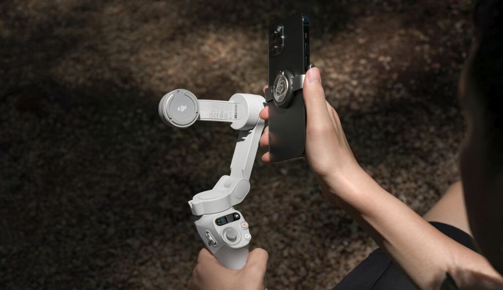 Osoba trzyma gimbal w kolorze białym, do którego przymocowany jest smartfon. Na gimbalu widoczne jest logo DJI.