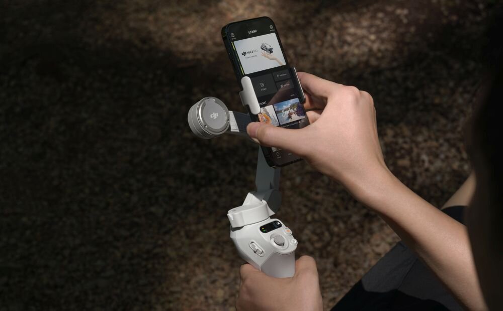 Osoba trzyma gimbal z zamocowanym smartfonem, na którym widoczny jest interfejs aplikacji. Na gimbalu znajduje się logo \'dji\'.